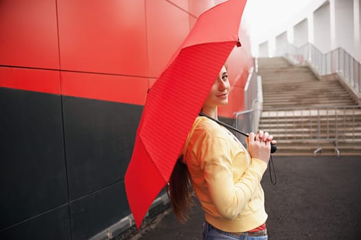 A pretty girl with long red hair, with a red umbrella in her hands, walks and smiles. Portrait in the open air