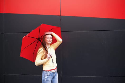 A pretty girl with long red hair, with a red umbrella in her hands, walks and smiles. Portrait in the open air