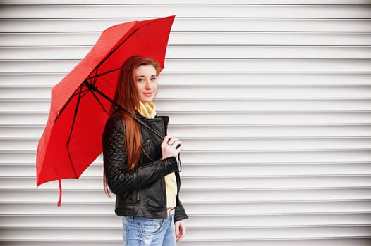 A pretty girl with long red hair, in a black leather jacket with a red umbrella in her hands, walks and smiles. Portrait in the open air