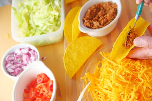 A man puts ground beef into a taco shell with grated cheese, shredded lettuce, chopped tomatoes and onions nearby