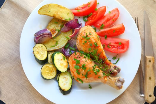 Roasted chicken legs with tomatoes, fingerling potatoes, onions, zucchini, rosemary and scallions on a white plate