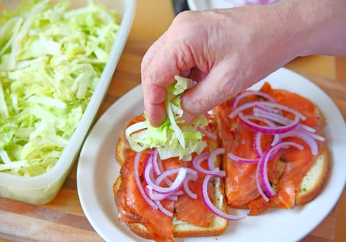 A man puts shredded lettuce on a smoked salmon sandwich