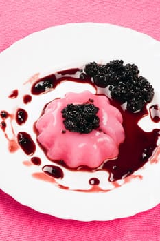 Pudding with sauce of blackberry is on white plate on pink background.