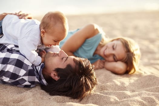 Baby boy laughing while playing with his dad on the beach while his mom smiles on in the background