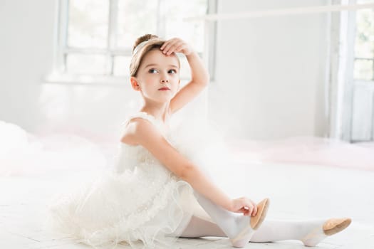 The little balerina in white tutu in class at the ballet school sitting on the wooden floor