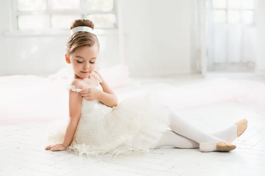 The little balerina in white tutu in class at the ballet school sitting on the wooden floor
