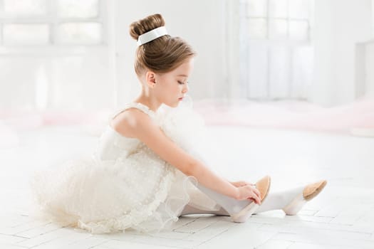The little balerina in white tutu in class at the ballet school sitting on the wooden floor