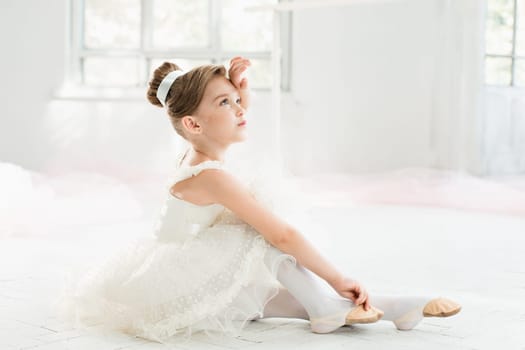 The little balerina in white tutu in class at the ballet school sitting on the wooden floor