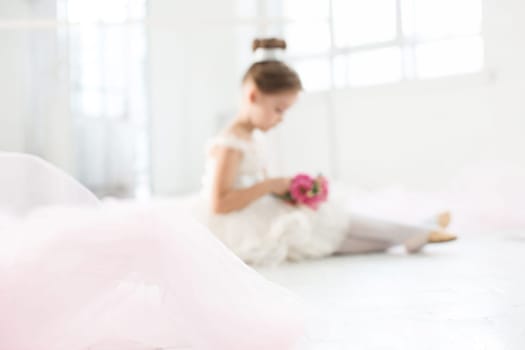 The little balerina in white tutu in class at the ballet school sitting on the wooden floor with flowers