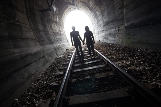 Couple walking hand in hand along the track through a railway tunnel towards the bright light at the other end, they appear as silhouettes against the light