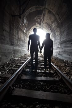 Couple walking hand in hand along the track through a railway tunnel towards the bright light at the other end, they appear as silhouettes against the light