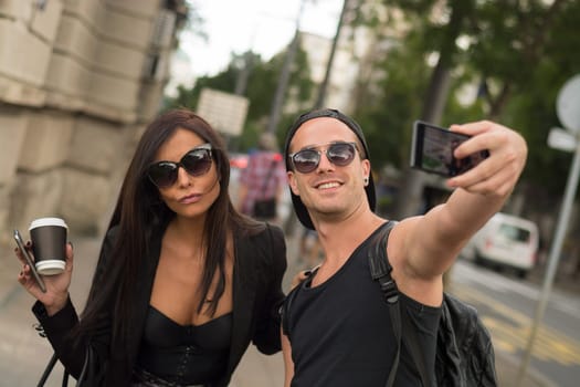 Two cheerful friends taking photos of themselves on smart phone, urban city outdoor scene, selective focus