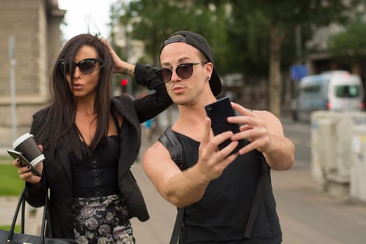Two cheerful friends taking photos of themselves on smart phone, urban city outdoor scene, selective focus