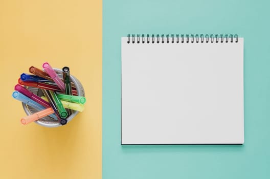 Office workplace minimal concept. Blank notebook, color pen box on yellow and green turquoise background. Top view with copy space, flat lay. Pastel color filter.