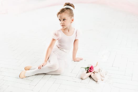 The little balerina in white tutu in class at the ballet school sitting on the wooden floor with flowers