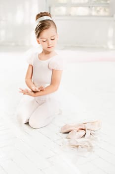 The little balerina in white tutu in class at the ballet school sitting on the wooden floor with flowers