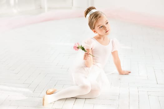 The little balerina in white tutu in class at the ballet school sitting on the wooden floor with flowers