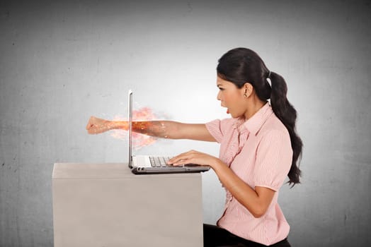 Furious asian business woman throws a punch into the laptop on grey background