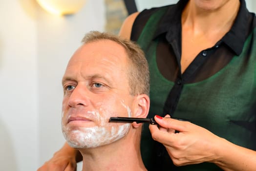 female hairdresser shaving beard of man in her shop