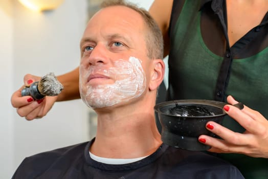 female hairdresser shaving beard of man in her shop