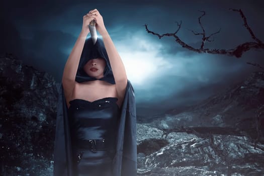Witch woman with black costume wearing cloak and holding knife on the rock hills at the midnight