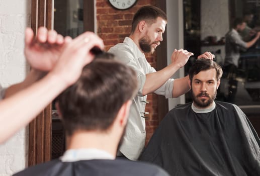 Young handsome barber making haircut of attractive bearded man in barbershop