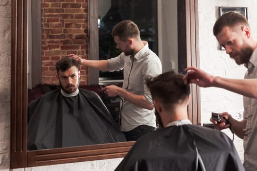 Young handsome barber making haircut of attractive bearded man in barbershop
