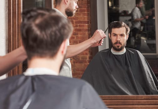 Young handsome barber making haircut of attractive bearded man in barbershop