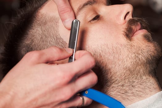 Hipster client visiting barber shop. The hands of young barber making the cut of beard