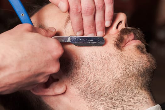 Hipster client visiting barber shop. The hands of young barber making the cut of beard