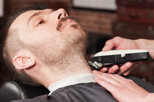 Hipster client visiting barber shop. The hands of young barber making the cut of beard