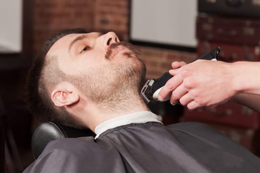 Hipster client visiting barber shop. The hands of young barber making the cut of beard