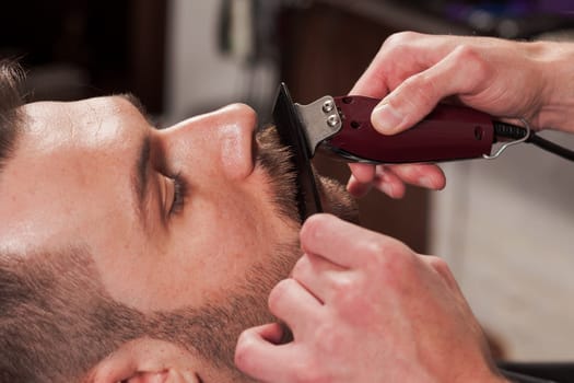 Hipster client visiting barber shop. The hands of young barber making the cut of beard