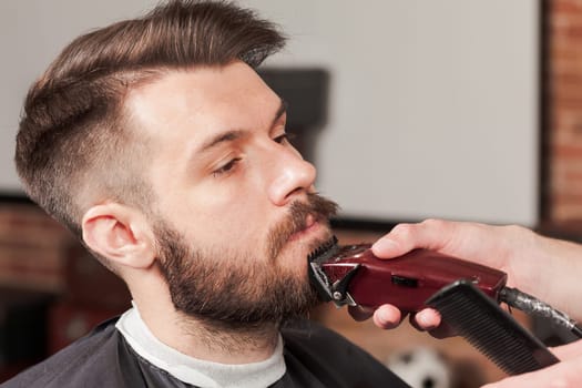 Hipster client visiting barber shop. The hands of young barber making the cut of beard