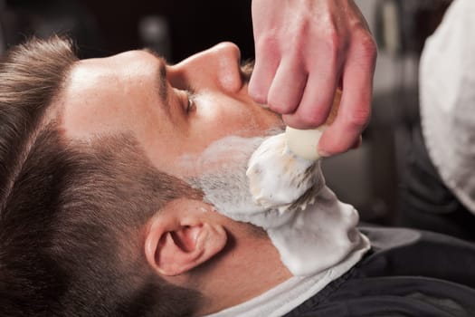 Hipster client visiting barber shop. The hands of young barber making the cut of beard
