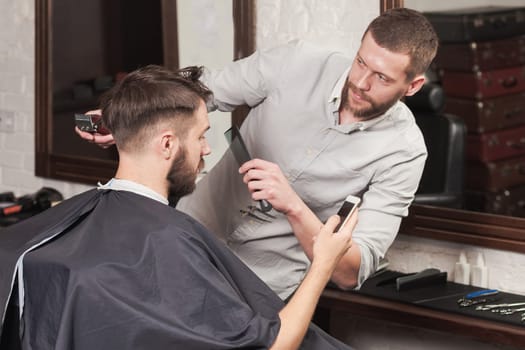 Young handsome barber making haircut of attractive bearded man in barbershop