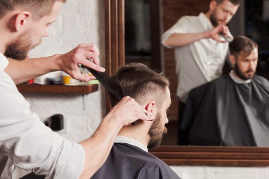 Young handsome barber making haircut of attractive bearded man in barbershop