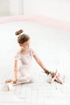 The little balerina in white tutu in class at the ballet school sitting on the wooden floor with flowers