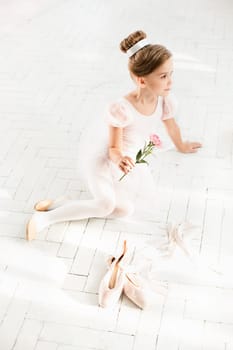 The little balerina in white tutu in class at the ballet school sitting on the wooden floor with flowers