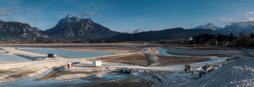Panoramic view of the drained Forggensee near Füsssen.. The Forggensee,also stored Roßhaupten,is a reservoir crossed by the Lech near Füssen in the Königswinkel in the Bavarian Allgäu. With the meltwater from the surrounding mountains,sedimentary rock and debris settle in the Forggensee,which is collected and transported in winter with drained water,by excavators,bulldozers and other heavy equipment.