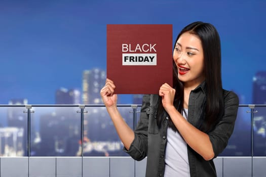 Happy asian woman holding board with Black Friday message over cityscape background