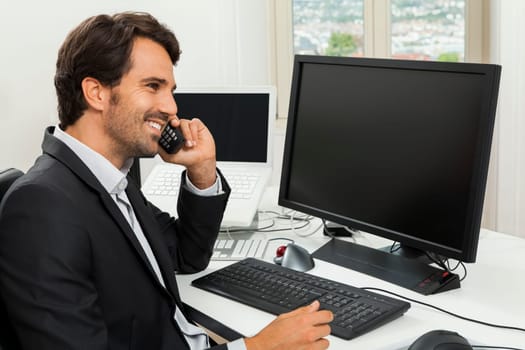 Successful businessman sits in the office and makes phone calls at his computer workplace in black suit