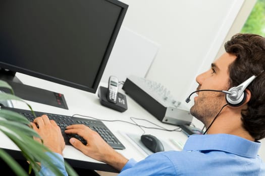 Attractive young man with headset in the office at customer service operator phone service cheerful with blue shirt