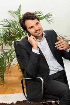 Successful businessman sits in the office and makes phone calls at his computer workplace in black suit