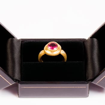 Golden ring with pink gem is in black jewelry box on white background.