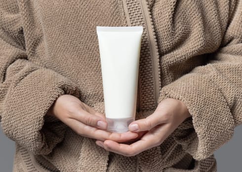 Close up of woman in light brown bathrobe with white blank cream tube in hands, mockup. Hands with natural manicure holding squeeze cosmetic tube - lotion or shampoo