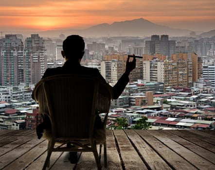 Silhouette of businessman sit on chair and hold a cigar and looking at the city in night.