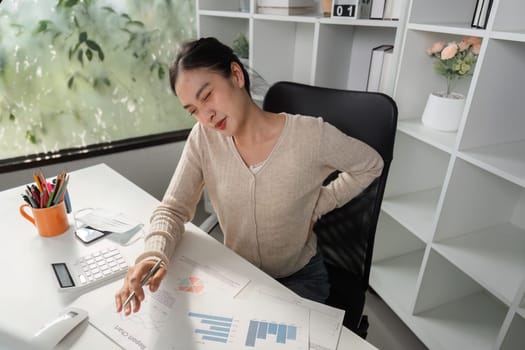 A woman feeling back pain at her desk, struggling to analyze financial charts and graphs in a contemporary office environment.