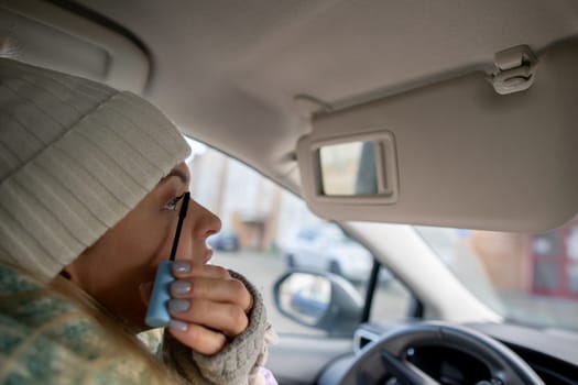 A stylish woman in a cozy beanie applies mascara in her car, showcasing her winter beauty routine and unique style