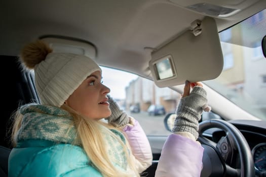 A fashionable woman dressed in a cozy winter outfit elegantly checks her appearance in the cars side mirror